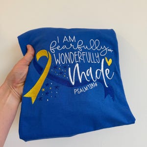 May include: Royal blue t-shirt with embroidered text "I AM fearfully + wonderfully MADE" and "PSALM 139:14". Features a yellow and blue ribbon design with star accents. The shirt is held by a hand.