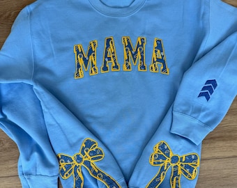May include: Light blue sweatshirt with the word "MAMA" in yellow and blue lettering across the chest. The sleeves feature yellow and blue bow designs and blue arrow details. A comfortable, casual top.