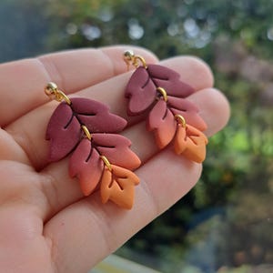 Op de afbeelding: Oorbellen met een bladvorm in herfsttinten. Elke oorbel heeft drie bladeren in bordeauxrood, oranje en geel, verbonden door goudkleurig metaal. De bladeren zijn in een trapsgewijze opstelling gerangschikt, waardoor een herfstthema-accessoire ontstaat.