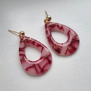 Op de afbeelding: Een paar druppelvormige oorbellen. De oorbellen hebben een doorschijnende roze basis met een gemarmerd bordeauxrood patroon en gouden spikkels. Elke oorbel heeft een goudkleurige pin en baldetail.
