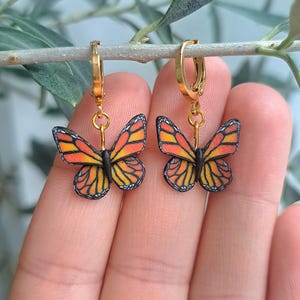 Op de afbeelding: Een paar goudkleurige oorringen met vlinderhangers. De vlinderhangers hebben oranje, gele en zwarte vleugels. De oorbellen hangen aan een goudkleurige ring. De achtergrond is wazig.