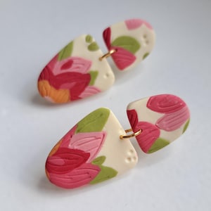 Op de afbeelding: Een paar handgemaakte polymeerklei oorbellen. Elke oorbel heeft een crèmekleurige, druppelvormige basis met een bloemenontwerp in roze, rode, oranje en groene tinten. De oorbellen zijn verbonden door een kleine goudkleurige ring.
