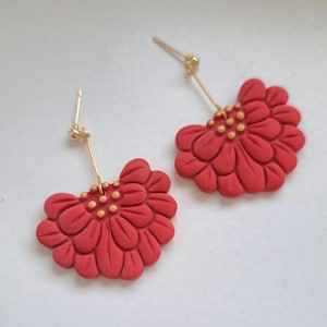 Op de afbeelding: Een paar rode bloemvormige oorbellen met goudkleurige accenten. De oorbellen hebben een stekersluiting en een goudkleurige draad die de bloem met de steker verbindt. De bloemblaadjes zijn gedetailleerd en getextureerd.