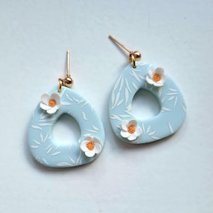 Op de afbeelding: Een paar lichtblauwe druppelvormige oorbellen met een wit bladerpatroon en kleine witte bloemen met oranje centra. De oorbellen hebben goudkleurige oorhaken en zijn een uniek accessoire.