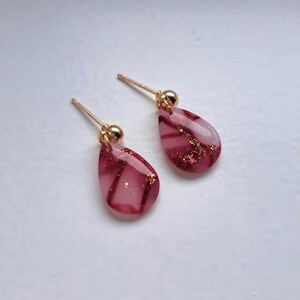 Op de afbeelding: Een paar druppelvormige oorbellen. De oorbellen hebben een doorschijnend roze en rood ontwerp met gouden accenten. De pinnen en kogelknopjes zijn goudkleurig. De oorbellen worden gepresenteerd op een wit oppervlak.