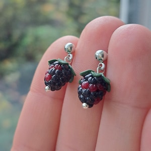 Op de afbeelding: Een paar oorhangers met miniatuur bramen. Elke oorbel heeft een zilverkleurige knop en een gedetailleerde braambedeltje met rode en zwarte kleuren en groene bladeren. De oorbellen zijn ontworpen om op de vrucht te lijken.