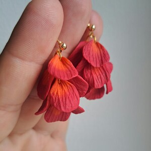 Op de afbeelding: Handgemaakte oorbellen met een trapsgewijs bloemendesign. De oorbellen tonen gelaagde bloemblaadjes in tinten rood en oranje, met goudkleurige oorhaken. De bloemblaadjes hebben een textuur, wat diepte aan het ontwerp toevoegt.
