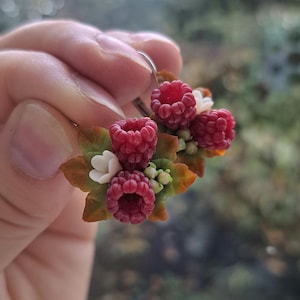 Könnte beinhalten: Handgefertigte Ohrringe mit Miniatur-Himbeeren, weißen Blüten und grünen und braunen Blättern. Die Ohrringe werden vor einem verschwommenen Hintergrund gehalten, der die filigranen Details des Polymer-Ton-Designs zeigt.