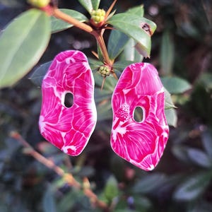 Op de afbeelding: Een paar roze en witte bloemen oorbellen met een druppelvorm en een klein gaatje. De oorbellen staan tegen een achtergrond van groene bladeren en takken, wat een natuurlijk contrast creëert.