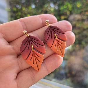 Op de afbeelding: Een paar oorringen in de vorm van bladeren met een kleurovergang van donkerrood naar oranje. De oorbellen zijn gemaakt van een glad materiaal en hebben goudkleurige oorbelpalen en -ringen. Het ontwerp lijkt op herfstbladeren.