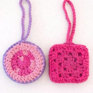 May include: Two handmade crochet ornaments. One is round with pink, light pink, and purple concentric circles. The other is square, in a solid pink color. Both have a loop for hanging.
