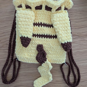 May include: A yellow and brown crochet drawstring backpack featuring two long yellow ears with brown tips, two horizontal brown stripes across the center, and a yellow and brown tail attached to the bottom. The bag has brown crochet straps.