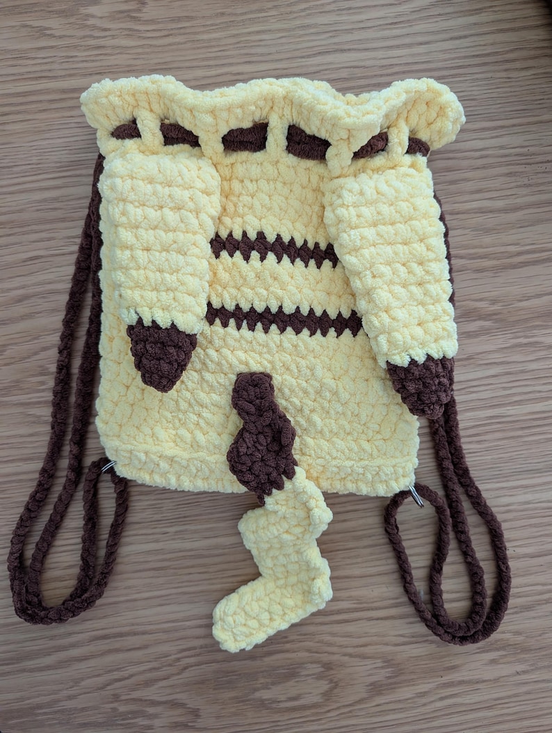 May include: A yellow and brown crochet drawstring backpack featuring two long yellow ears with brown tips, two horizontal brown stripes across the center, and a yellow and brown tail attached to the bottom. The bag has brown crochet straps.
