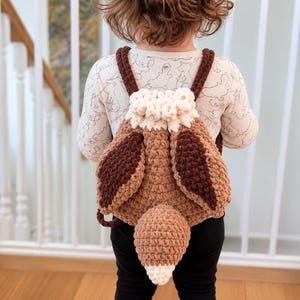 May include: A crochet backpack designed to resemble a fox, featuring a light brown body, dark brown ear-like flaps, a white drawstring top, and a prominent fox tail attached to the center. The bag has two dark brown straps for carrying.