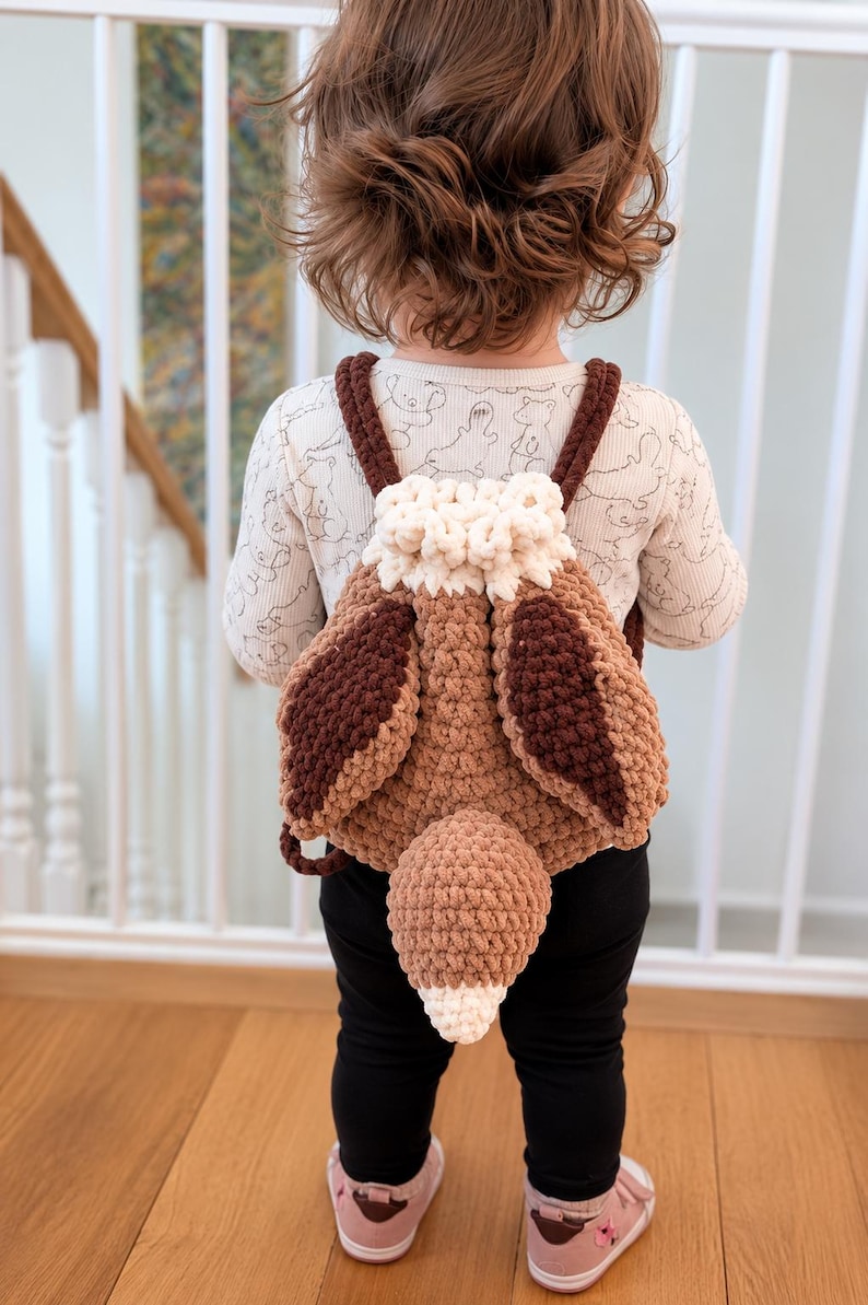 May include: A crochet backpack designed to resemble a fox, featuring a light brown body, dark brown ear-like flaps, a white drawstring top, and a prominent fox tail attached to the center. The bag has two dark brown straps for carrying.