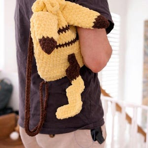 May include: A yellow and brown crocheted drawstring backpack shaped like a creature with pointed ears, dark brown tips, and a jagged tail. The bag features a soft, textured yarn construction with long, thin brown crocheted straps for carrying.
