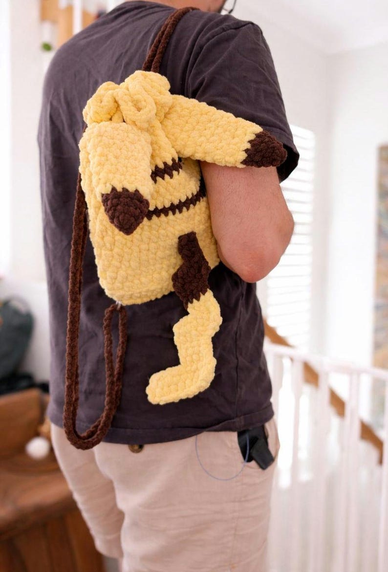 May include: A yellow and brown crocheted drawstring backpack shaped like a creature with pointed ears, dark brown tips, and a jagged tail. The bag features a soft, textured yarn construction with long, thin brown crocheted straps for carrying.