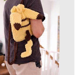 May include: A yellow and brown crocheted drawstring backpack featuring a character design with long ears, a jagged tail, and brown accents on the tips. The bag is constructed from thick, soft yarn and includes two brown crocheted shoulder straps.