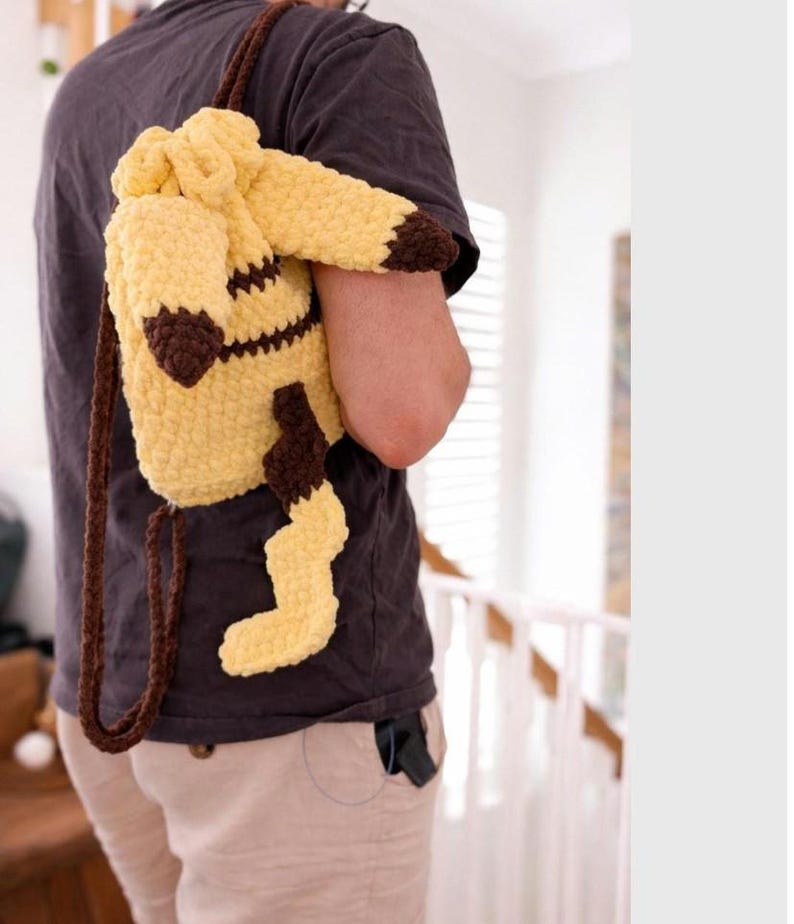 May include: A yellow and brown crocheted drawstring backpack featuring a character design with long ears, a jagged tail, and brown accents on the tips. The bag is constructed from thick, soft yarn and includes two brown crocheted shoulder straps.
