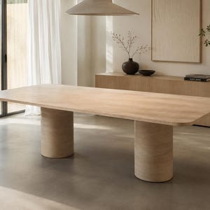 Puede incluir: Una mesa de comedor rectangular grande con acabado de veta de madera clara, con esquinas redondeadas y dos bases de pedestal cilíndricas sustanciales. La superficie de la mesa es lisa y plana, apoyada por dos patas gruesas y sólidas.