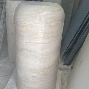 Op de afbeelding: Een cilindervormig voetstuk van beige travertijnsteen met een gladde, gepolijste afwerking en zichtbare horizontale natuurlijke aders. De structuur heeft een afgeronde bovenrand en een solide, stevige basis die de textuur toont.