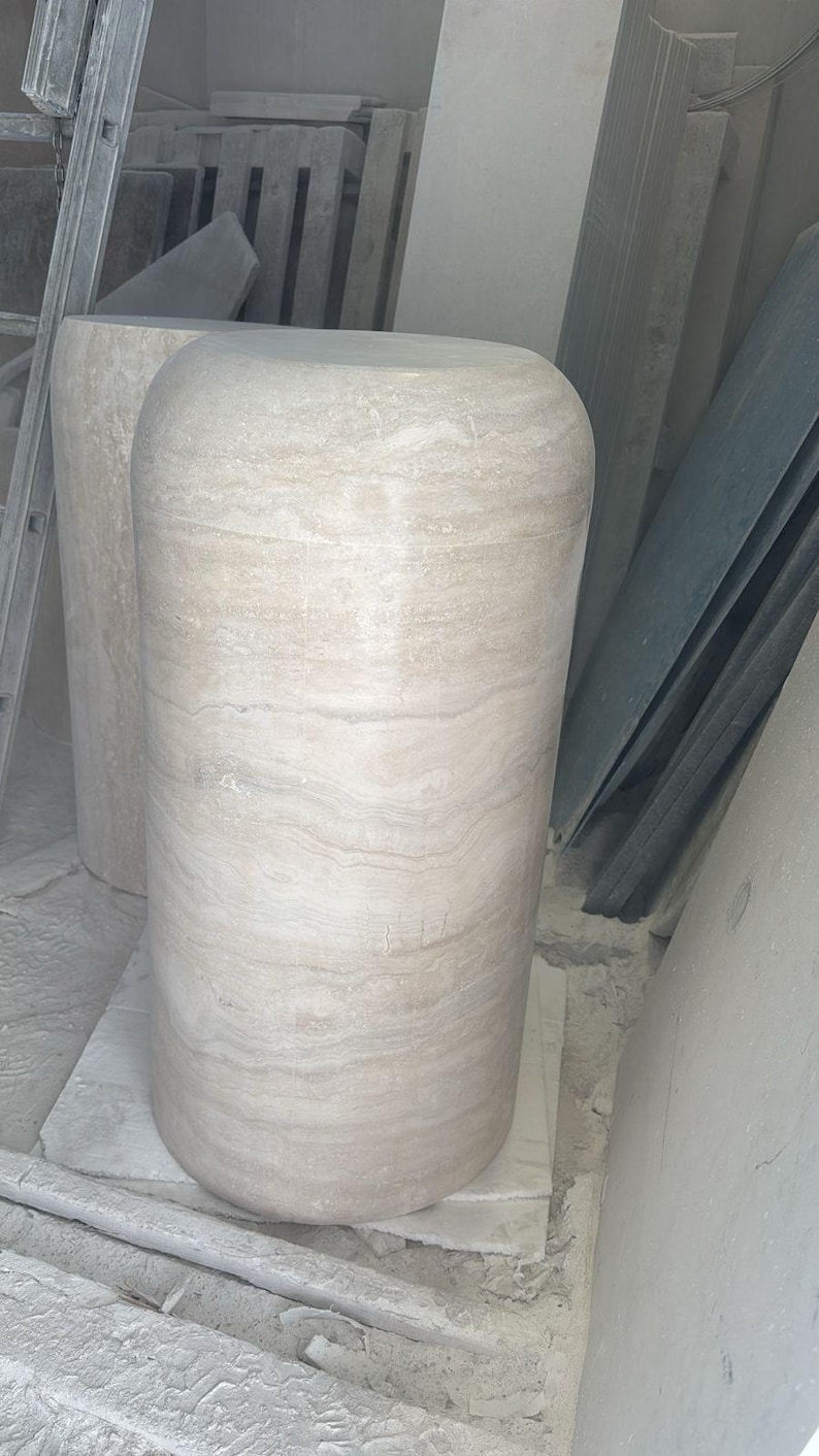 Op de afbeelding: Een cilindervormig voetstuk van beige travertijnsteen met een gladde, gepolijste afwerking en zichtbare horizontale natuurlijke aders. De structuur heeft een afgeronde bovenrand en een solide, stevige basis die de textuur toont.