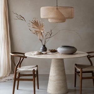 May include: A round beige stone dining table with a conical base, accompanied by two wooden chairs with woven seats. Above hangs a tiered cylindrical woven pendant light. On the table sits a dark gray vase with dried foliage and a gray bowl.