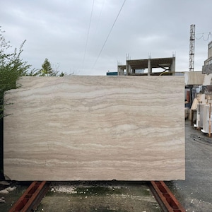 Op de afbeelding: Een grote, rechthoekige plaat van beige travertijnsteen met opvallende horizontale aders en natuurlijke gelaagde patronen. Het oppervlak heeft een gladde, gepolijste afwerking met subtiele variaties in lichtbruine en cr&egrave;mekleurige tinten.