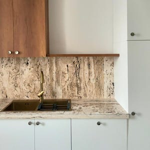 Puede incluir: Un diseño de cocina moderno con un grifo y fregadero de latón sobre un salpicadero texturizado. Armarios blancos con pomos oscuros y armarios superiores de madera completan el conjunto. La encimera tiene un patrón claro y moteado.