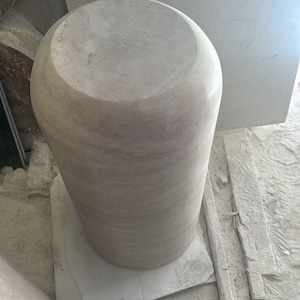 Op de afbeelding: Een cilindervormig stenen voetstuk met een glad, lichtbeige oppervlak en subtiele horizontale aders. Het object heeft een platte, ronde bovenkant en een massief, afgerond lichaam met een natuurlijke, onbewerkte textuur.