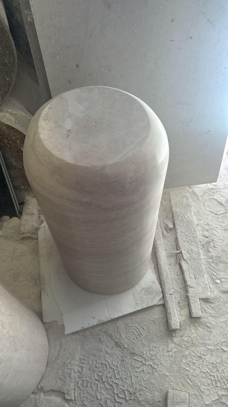 Op de afbeelding: Een cilindervormig stenen voetstuk met een glad, lichtbeige oppervlak en subtiele horizontale aders. Het object heeft een platte, ronde bovenkant en een massief, afgerond lichaam met een natuurlijke, onbewerkte textuur.