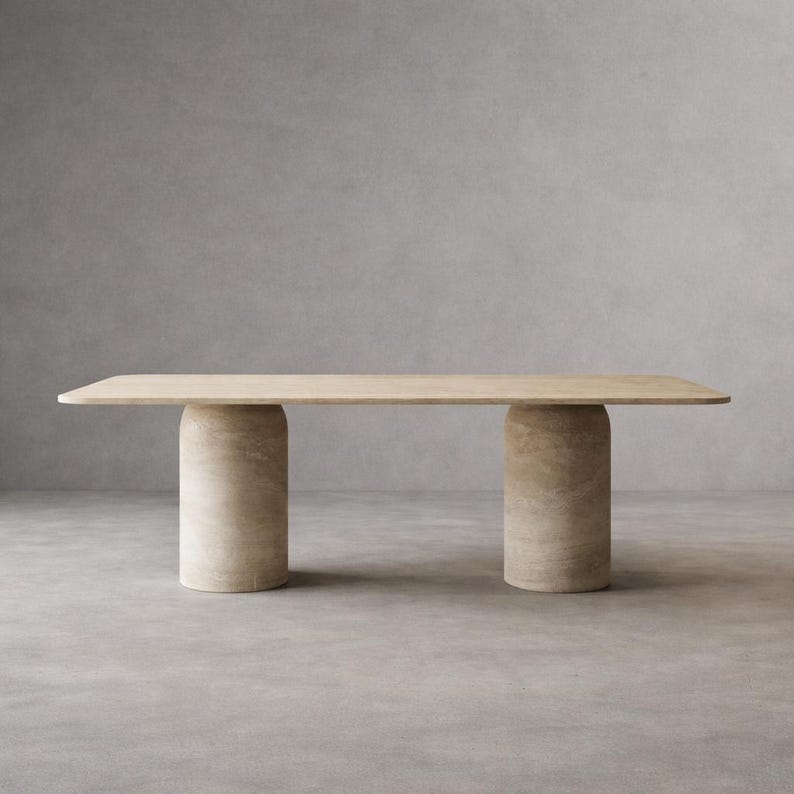 Op de afbeelding: Een rechthoekige eettafel met een glad, lichtbeige stenen oppervlak met afgeronde hoeken. De tafel wordt ondersteund door twee dikke, cilindrische stenen zuilen die qua kleur en textuur bij het tafelblad passen, wat een solide structuur vormt.