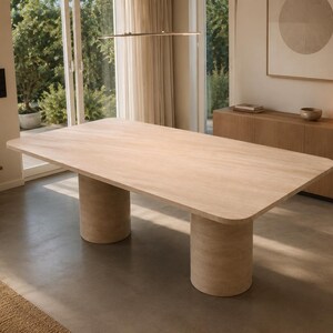May include: A large rectangular dining table with a light wood grain finish and rounded corners. The table is supported by two substantial cylindrical pedestal bases that match the tabletop, creating a cohesive and stable furniture piece.