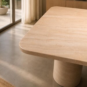 May include: A rectangular dining table with a smooth, light beige travertine stone surface featuring natural horizontal veining and rounded corners. The table is supported by a single, thick, cylindrical pedestal base made of the same stone.