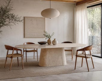 May include: An oval dining table with a thick, light-colored stone pedestal base and a smooth top. Four wooden chairs with woven seats surround the table. On the table sit a dark bowl, a light-colored vase, and a smaller rounded vessel.