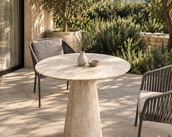 May include: A round beige stone table with a conical pedestal base, accompanied by two dark gray chairs featuring woven backrests and light gray seat cushions. A small beige ceramic vase and a matching bowl sit on the tabletop surface.