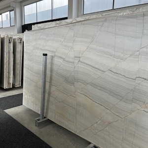 Peut inclure: Une grande dalle rectangulaire en pierre claire présentant des veines horizontales marquées dans des tons gris, blancs et beige subtil. La surface affiche un motif naturel fracturé avec de fines lignes sombres traversant la face.