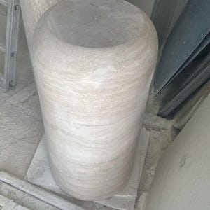 Op de afbeelding: Een cilindervormig stenen voetstuk met een gladde, afgeronde bovenkant en een lichtbeige, gemarmerd oppervlak met horizontale aders. De structuur heeft een solide, zwaar uiterlijk met een consistente textuur over het hele lichaam.