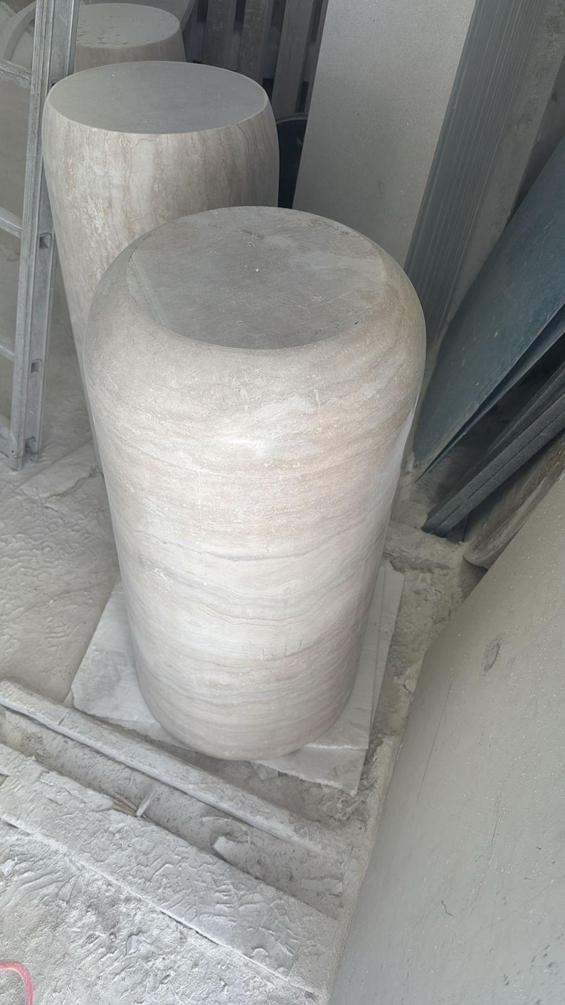 Op de afbeelding: Een cilindervormig stenen voetstuk met een gladde, afgeronde bovenkant en een lichtbeige, gemarmerd oppervlak met horizontale aders. De structuur heeft een solide, zwaar uiterlijk met een consistente textuur over het hele lichaam.