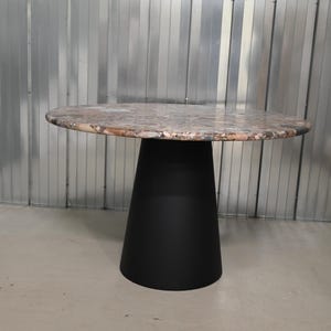Op de afbeelding: Een ronde tafel met een gespikkelde, veelkleurige bovenkant en een zwarte, conische voet. Het tafelblad lijkt van steen of een vergelijkbaar materiaal te zijn. De voet is effen, matzwart. Deze tafel is geschikt voor een eetkamer of woonkamer.