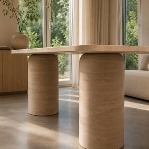 May include: A dining table featuring a rectangular tabletop with rounded corners, supported by two large, cylindrical pedestal bases. The entire structure is finished in a light beige, textured stone material with visible natural veining.