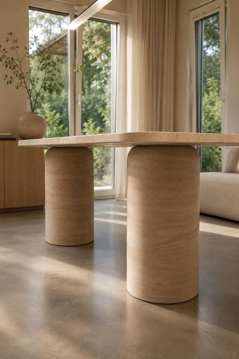 May include: A dining table featuring a rectangular tabletop with rounded corners, supported by two large, cylindrical pedestal bases. The entire structure is finished in a light beige, textured stone material with visible natural veining.