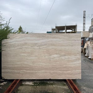 May include: A large, rectangular slab of beige travertine stone featuring horizontal, wavy, cream and tan veining patterns. The natural stone slab has a smooth, polished surface and raw, unfinished edges, standing upright on a metal support frame.