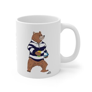 Puede incluir: Una taza de cerámica blanca con una ilustración de dibujos animados de un oso pardo que viste una camiseta de rugby a rayas azul marino y blancas. El oso sostiene una lata de bebida amarilla y un balón de rugby azul y blanco bajo el brazo.