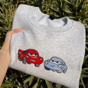Puede incluir: Una sudadera gris claro con im&aacute;genes bordadas de dos coches de dibujos animados: un coche rojo con el n&uacute;mero 95 y un coche azul. La sudadera est&aacute; hecha de un material suave y tiene cuello redondo.