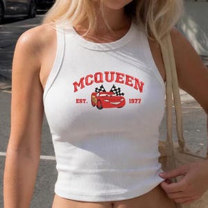 Puede incluir: Camiseta sin mangas blanca acanalada con la palabra "McQueen" en letras rojas arqueadas sobre un coche de dibujos animados rojo con banderas a cuadros. El texto "EST. 1977" est&aacute; debajo del coche. La camiseta sin mangas es sin mangas.