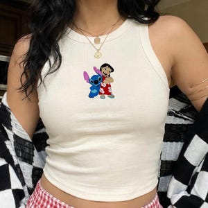 May include: Cream-colored tank top featuring a colorful graphic of Stitch and Lilo from the Disney movie Lilo & Stitch. The top is a ribbed style and is paired with red and white checkered pajama pants. A black and white checkered cardigan is worn over the top.