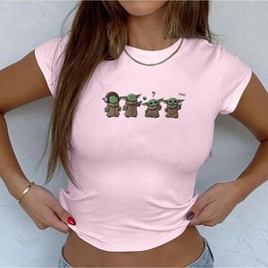 May include: Light pink t-shirt featuring a graphic of four Grogu characters in various poses. One Grogu wears headphones, another holds a ball, and a third has a question mark above its head. The word "cool" is written next to the fourth Grogu.
