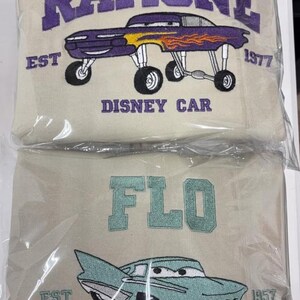 May include: Two beige sweatshirts with embroidered designs. The top sweatshirt features the word "RAMONE" in purple, a purple car with flames, and the text "DISNEY CAR" and "EST 1977". The bottom sweatshirt says "FLO" and has a teal car with the text "EST 1957".