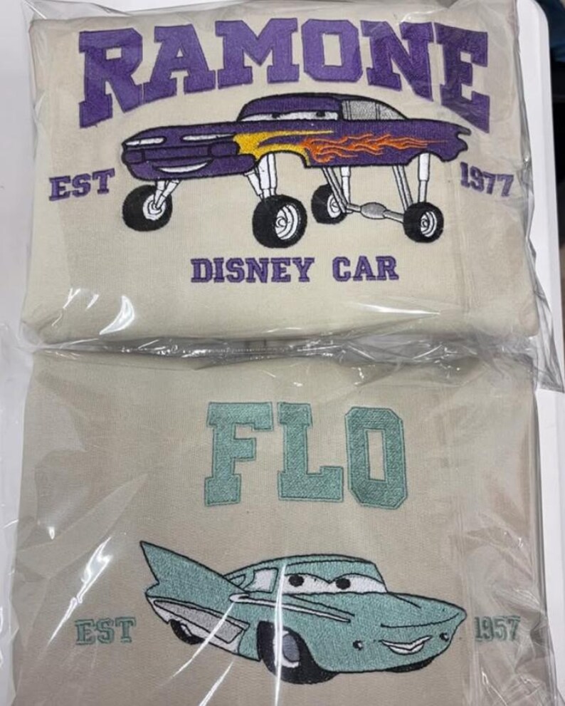 May include: Two beige sweatshirts with embroidered designs. The top sweatshirt features the word "RAMONE" in purple, a purple car with flames, and the text "DISNEY CAR" and "EST 1977". The bottom sweatshirt says "FLO" and has a teal car with the text "EST 1957".
